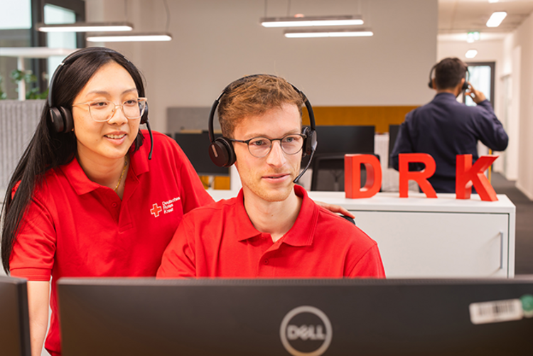 Imagebild rotkreuzCRM | Kunden Dieses Bild zeigt zwei Mitarbeitende des DRK in DRK-Poloshirts mit Headsets auf dem Kopf, die gemeinsam auf einen Monitor gucken.