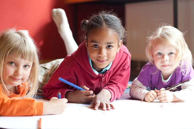 rotkreuzCRM | Familie Dieses Imagebild zeigt drei Kinder, die auf dem Boden liegen, malen und direkt in die Kamera gucken.