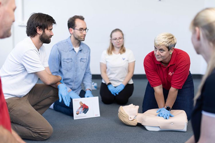 rotkreuzCRM | Kurse Dieses Bild zeigt eine Schulungssituation einer Rotkreuzkurses für Erste Hilfe. Die Kursleiterin demonstriert den teilnehmenden die Herzdruckmassage an einer Übungspuppe.